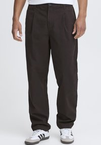 Pantaloni marrone scuro, dalla vestibilità ampia, realizzati in cotone con pieghe frontali, zip nascosta e tasche laterali, abbinati a scarpe sportive bianche e grigie.