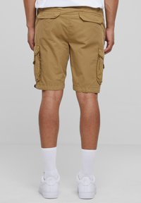 Beige cargoshorts met zij- en achterzakken, katoenen stof, met zoom afgewerkte randen, gedragen met witte sokken en sneakers. Effen achtergrond.