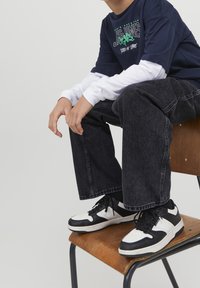 Marinblå graphic t-shirt lager på en vit långärmad skjorta, parat med svarta vida jeans och svart-vita sneakers på en trästol.