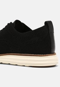 Svart mesh-sneaker med en texturerad ovandel, läderdetalj vid hälen och en vit flerskikts-sula med en brun mellansula.