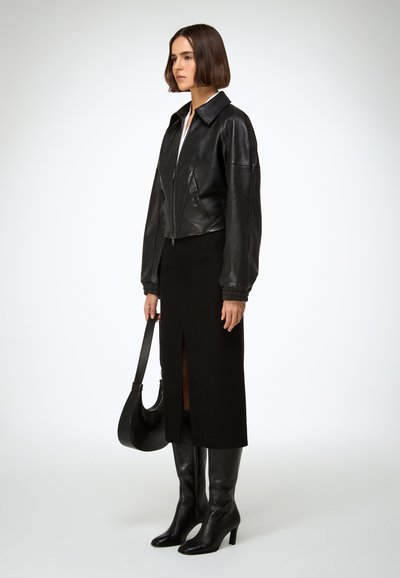 Crna kožna skraćena jakna s patentnim zatvaračem, crna midi suknja s bočnim prorezom i crne čizme do koljena. Modni dodaci uključuju crnu torbicu za rame.