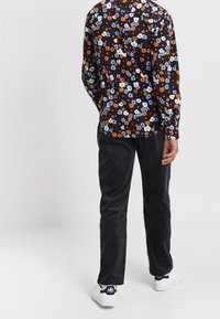 Camisa negra con estampado floral en azul, naranja y blanco; combinada con pantalones oscuros de pana y zapatillas blancas.
