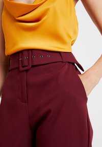 Yellow satin cowl neck top paired with high-waisted maroon trousers featuring a belt, silver hardware, and front pockets.