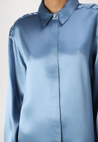 Camicia di satin azzurro chiaro con colletto classico, maniche lunghe e patta con bottoni frontale; presenta una texture liscia e lucida con una leggera lucentezza.