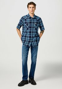 Korte mouwen blauwe geruite overhemd met donkerblauwe en groene accenten, knoopsluiting kraag en voorzakken, gecombineerd met blauwe spijkerbroek en zwarte schoenen.