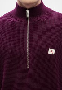 Πουλόβερ σε σκούρο μωβ με υφασμένο ανάγλυφο σχέδιο, ψηλό γιακά και ασημένιο φερμουάρ, με λευκή ετικέτα που φέρει κόκκινη λεπτομέρεια λογότυπου.