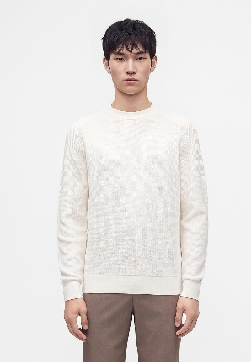 Jeune homme aux cheveux courts noirs se tenant face à l'avant, portant un pull blanc texturé à manches longues et un pantalon marron sur un fond uni.