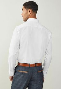 Camisa blanca de manga larga con un cuello clásico, combinada con pantalones a cuadros que tienen acentos marrones y un cinturón de cuero marrón.