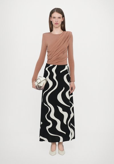 Jupe longue noire avec des motifs ondulés blancs associée à un haut drapé à manches longues marron. Le mannequin tient un petit sac à main en argent. Talons blancs.