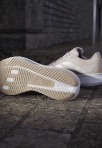 Baskets de sport beige clair avec une texture sur le dessus, des accents blancs et une semelle en caoutchouc présentant un motif adhérent et un détail de logo.
