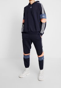 Marinblå hoodie med vita ränder, orange och blå ärmuppvakt, i kombination med matchande marinblå joggers med färgblockerade ränder.