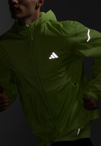 Neon-grüne, leichte Jacke mit Reißverschluss vorne, Logo und reflektierenden Akzenten auf der Brust und den Ärmeln; mit verstellbaren Manschetten und Saum.