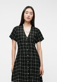 Čierne šaty s bielou mriežkovanou vzorom, krátkymi rukávmi, V-výstrihom a zlatými gombíkmi po celej dĺžke. Textúrovaná látka pridáva hĺbku.