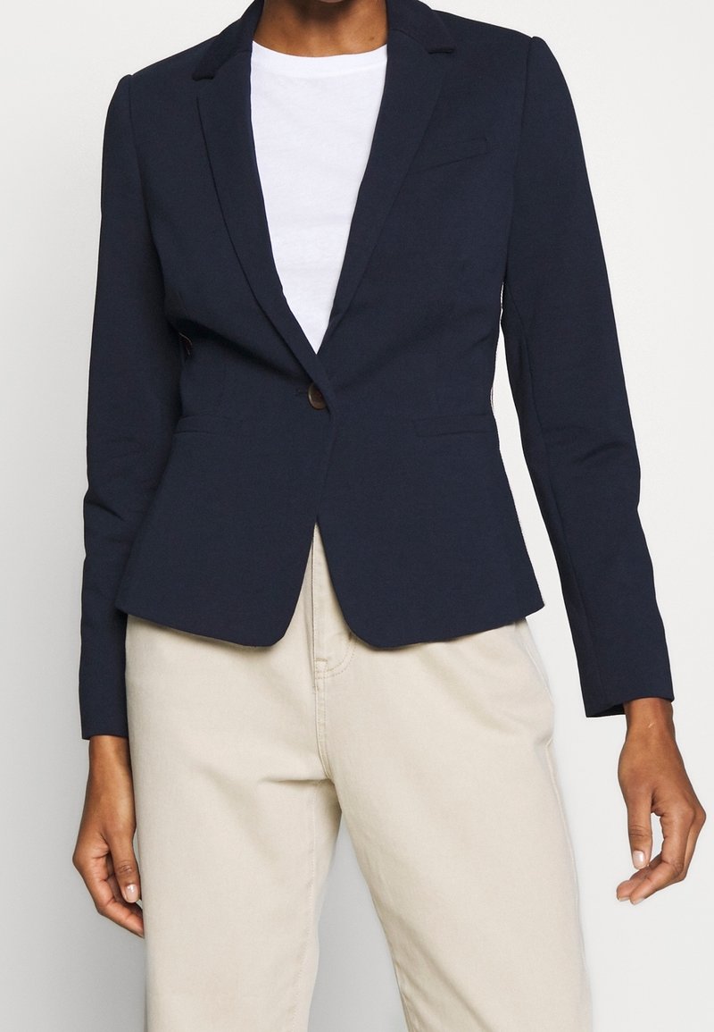 Personne portant un blazer bleu marine sur mesure par-dessus une chemise blanche et un pantalon beige, debout avec les bras relâchés le long du corps sur un fond uni.