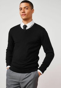 Suéter negro de cuello en V hecho de una tela suave, con mangas largas, puños acanalados y un diseño ajustado, utilizado sobre una camisa blanca con cuello.