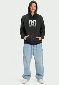 Sweatshirt com capuz preto com um logótipo DC em destaque à frente, combinado com jeans baggy azul claro e ténis brancos.