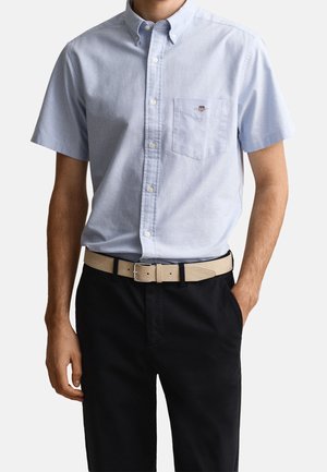 Homme portant une chemise bleu clair à manches courtes avec une poche poitrine et une ceinture beige avec un pantalon noir, une main dans la poche.