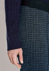 Linkerhand ontspannen naast gepatineerde blauw en zwart geblokte stoffen broek, met een mouw van een donkerpaarse gebreide trui zichtbaar.