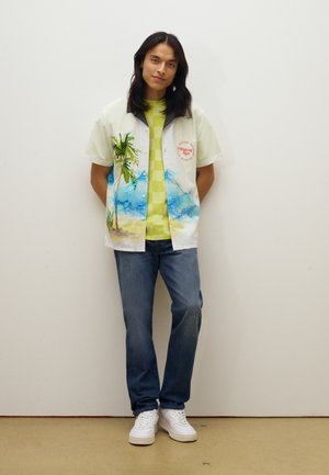 Korte mouwen, licht shirt met een palmboom- en strandprint, gelaagd over een groen geruit T-shirt, gecombineerd met blauwe jeans en witte sneakers.