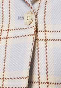 Gros plan d’un tissu à carreaux beige et bleu clair avec des lignes marron, montrant un bouton métallique rond brillant et une boutonnière.