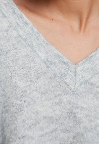 Hellgrauer V-Ausschnitt-Pullover mit weichem, strukturiertem Strick. Verfügt über ein dezentes marmoriertes Muster und gerippte Kanten am Ausschnitt.