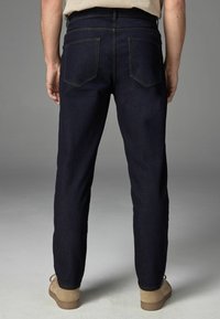Vaqueros de denim azul oscuro con corte recto, que presentan dos bolsillos traseros y detalles de costura sutiles. Combinados con zapatos de ante marrón claro.