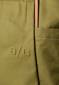 Olive green fabric bag with a textured surface, featuring a pink zipper and a side pocket. Engraved "a/u" logo on the front.