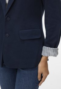 Marinblå blazer med två knappar, enkel bröstficka och randig manschett som syns från ärmen. Tyget verkar slätt och strukturerat; figursydd design.