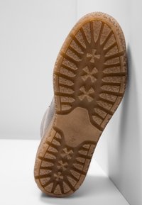 Brązowa gumowa podeszwa z teksturowanym bieżnikiem, z falistym wzorem i detalem logo. Cholewka ze zamszu w jasnoszarym kolorze, ustawiona pod kątem na białym tle.