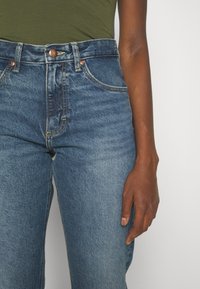 Hoge taille blauwe denim jeans met een vervaagde textuur, koperen knoopdetail en gestikte zakken, gecombineerd met een groene top.