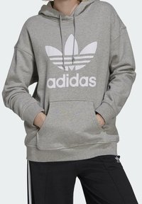 Grå huvtröja med vit Adidas-logotyp och tre-strimmigt mönster. Har framficka och dragsko i huvan. Mjuk tygtextur.