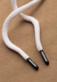 Bílé bavlněné tkaničky s černými plastovými špičkami, na kterých je nápis "Dombare", ležící na texturovaném béžovém pozadí.