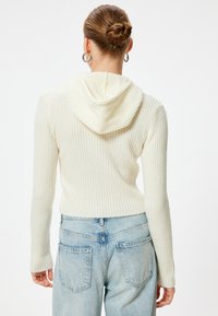 Cremfarbener gerippter Hoodie mit doppelter Kapuze und langen Ärmeln, kombiniert mit hellblauen Jeans. Weicher Stoff mit taillierter Silhouette.