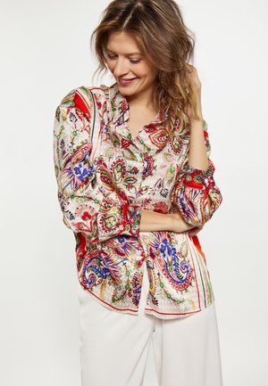 Lächelnde Frau mit welligem Haar trägt eine bunte Paisley-Bluse und weiße Hose, posiert mit einer Hand, die ihr Haar berührt.