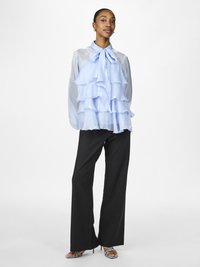 Helle blaue Chiffonbluse mit Rüschenschichten und einem Schleifenkragen, kombiniert mit schwarzen weiten Hosen und gemusterten hochhackigen Sandalen.