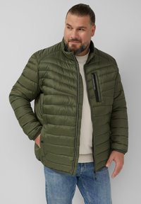 Padded Jacket aus olivgrünem Stoff mit gesteppter Optik, hohem Kragen, Zip-Verschluss und einer einzelnen Brusttasche mit schwarzem Akzent.