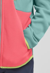 Giacca texture con un mix di pannelli rosa e teal, rifinita con un bordo verde lime. Il tessuto presenta un motivo a nido d'ape e polsini elastici.