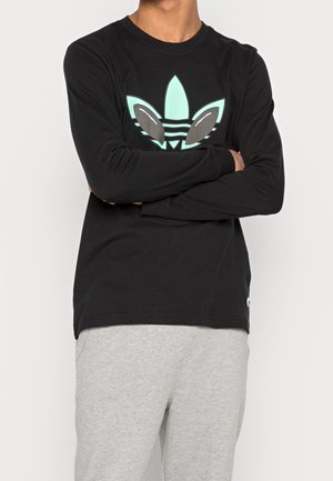 Osoba w czarnej koszulce z długim rękawem z zielono-szarym stylizowanym liściastym logo oraz jasnoszarych spodniach dresowych, z założonymi skrzyżowanymi ramionami.