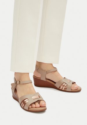 Béžové klínové sandály s texturovanou podrážkou, vybavené páskem na kotníku se sponou a kovovými doplňky na nártu, zdůrazňujícími vystřižený design.