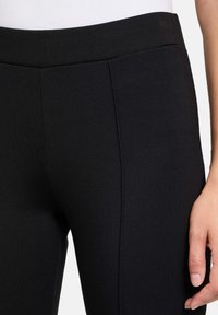 Leggings noirs en tissu extensible, avec une texture lisse et une coupe ajustée. Comprend des coutures verticales sur les côtés.