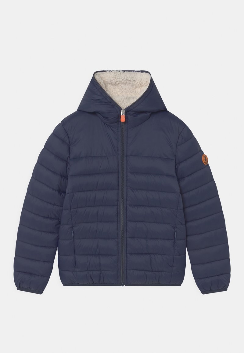Veste matelassée bleu marine avec capuche doublée en polaire blanche, fermeture éclair à l'avant, poches latérales et petite écusson rond marron sur la manche gauche.