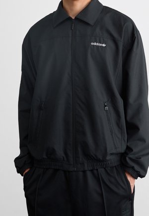 Mand iført sort Adidas lynlåstrøje med krave og lynlåslommer i siden, hænder i lommerne, mod en ensartet lys baggrund.