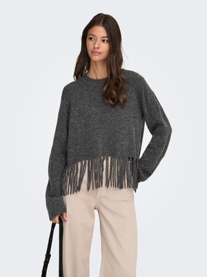 Femeie cu păr lung și castaniu, purtând un pulover gri închis cu franjuri și pantaloni bej deschis, ținând o geantă neagră de umăr într-o mână.