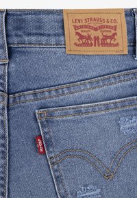 Niebieskie dżinsy z denimu z skórzaną metką, pomarańczowym przeszyciem, detalami na tylnej kieszeni oraz małą czerwoną metką z klasycznym wzornictwem.