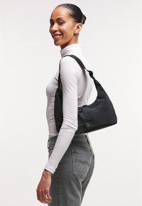 Borsa a spalla in tessuto nero con design curvo, dotata di chiusura a zip e tracolla regolabile. Il modello indossa un dolcevita bianco e jeans grigi.