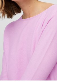 Jersey de color rosa claro con un patrón de tejido texturizado, que presenta un escote ancho y mangas largas, visto desde el hombro hacia arriba.