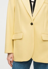 Άτομο που φοράει ανοιχτό κίτρινο σακάκι μονόκουμπο με πλαϊνές τσέπες πάνω από μπλε τζιν και μαύρο-άσπρο τοπ.