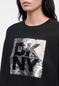 Femme portant un sweat-shirt noir à col rond avec un design carré en sequins argentés et noirs épelant "DKNY" sur la poitrine.