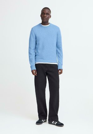 Jauns vīrietis valkā gaiši zilu džemperi, balu apakšveļu, melnas platas bikses un melnas sporta kurpes ar baltām svītrām, stāvot uz balta fona.