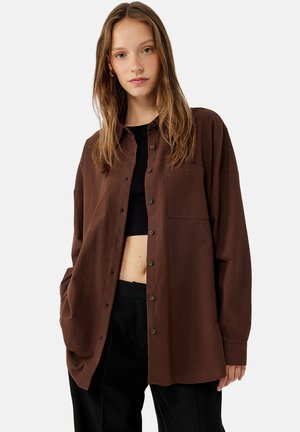 Chemise marron à boutons avec une poche, manches longues, coupe décontractée et texture douce, associée à un top noir court et un pantalon noir.